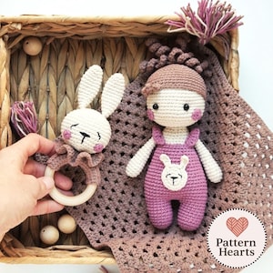 May include: Handmade crochet baby toys: a bunny rattle with a wooden ring and a doll in pink dungarees with a bunny motif. The toys are on a crocheted blanket and in a woven basket.
