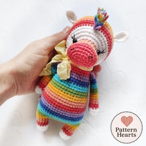 Puede incluir: Muñeca de unicornio de ganchillo hecha a mano, a rayas arcoíris, con un lazo de cinta amarilla. El unicornio tiene un hocico rosa, orejas blancas y una melena colorida. La muñeca se sostiene en una mano sobre un fondo blanco. La muñeca mide unos 15 cm de alto.