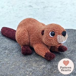 May include: A handmade plush beaver toy in shades of brown and burgundy. The beaver has large, black and white eyes, a beige muzzle, and a burgundy tail and feet. The toy is lying on a gray stone surface.