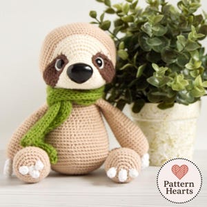 Könnte beinhalten: Eine gehäkelte Faultierpuppe mit beigem Körper, braunen Akzenten und einem grünen Schal. Das Faultier hat eine schwarze Nase und weiße Krallen. Eine Topfpflanze befindet sich im Hintergrund. Die Puppe ist handgefertigt.