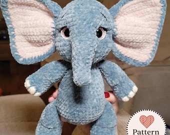 Può includere: Un peluche a forma di elefante realizzato in filato blu effetto velluto, con grandi orecchie dai pannelli interni rosa chiaro, occhi scuri con lunghe ciglia e dettagli bianchi sui piedi. È visibile un logo circolare con il testo Pattern Hearts.