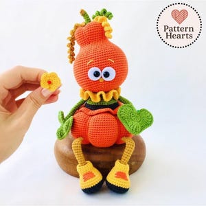 May include: A handmade orange crochet doll with large googly eyes, a yellow ruffled collar, and green leaves. The doll has yellow and black shoes and a green heart-shaped leaf. A small yellow flower is held nearby. The doll is sitting on a wooden base.