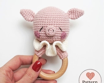 Può includere: Un sonaglio a forma di maialino all'uncinetto con testa in filato rosa chiaro, occhi chiusi ricamati, muso e guance rosa. Il maialino indossa un colletto bianco a balze ed è fissato a un anello in legno chiaro, liscio e rotondo.