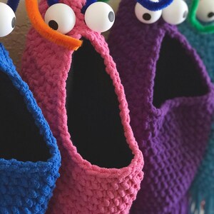 May include: A row of colorful, handmade monster-shaped storage baskets. Each basket is a different color, including blue, pink, purple, and teal, with large googly eyes and pipe cleaner antennae. The baskets have a wide opening and a black interior.