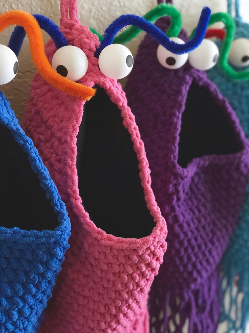 May include: A row of colorful, handmade monster-shaped storage baskets. Each basket is a different color, including blue, pink, purple, and teal, with large googly eyes and pipe cleaner antennae. The baskets have a wide opening and a black interior.