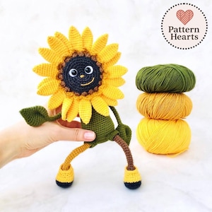 Puede incluir: Muñeca de girasol de ganchillo hecha a mano con una cara sonriente, pétalos amarillos y un centro oscuro. La muñeca tiene hojas, piernas y brazos verdes. Tres ovillos de lana amarillos y verdes están apilados junto a la muñeca. El texto "Pattern Hearts" está en un círculo.