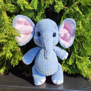 Op de afbeelding: Een handgemaakte, lichtblauwe gehaakte olifant knuffel met grote, roze gevoerde oren en blauwe ogen. De olifant heeft een lange slurf, kleine armen en benen en witte accenten op de voeten. Het speelgoed staat tegen een achtergrond van groen gebladerte.