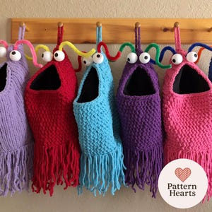 Może przedstawiać: Rząd kolorowych, ręcznie robionych toreb do przechowywania w kształcie potworów. Każda torba jest w innym kolorze, w tym fioletowym, czerwonym, niebieskim i różowym, z dużymi oczami i frędzlami. Torby wiszą na drewnianym wieszaku.