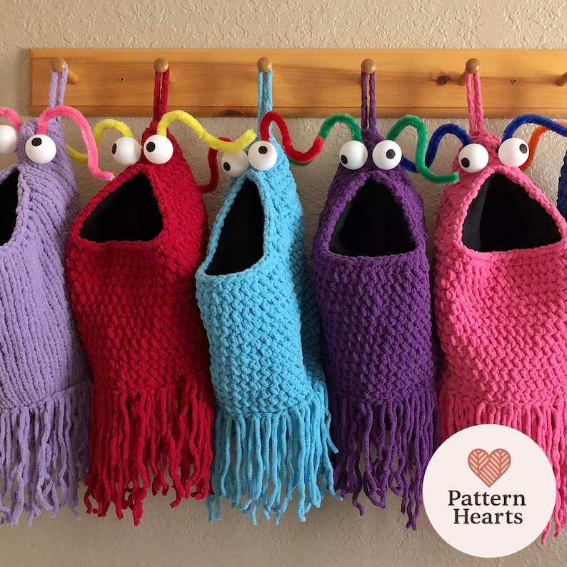 May include: A row of colorful, handmade monster-shaped storage bags. Each bag is a different color, including purple, red, blue, and pink, with large googly eyes and fringe. The bags hang from a wooden rack.