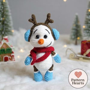 Puede incluir: Un muñeco de nieve de ganchillo con cuerpo blanco, que lleva una bufanda roja, orejeras azules y guantes y botas azules. El muñeco de nieve tiene una nariz de zanahoria y cuernos marrones. La imagen incluye un logotipo que dice "Pattern Hearts".