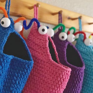 May include: A row of colorful, handmade crochet monster-shaped storage bags hanging on a wooden rack. Each bag is a different color: blue, pink, purple, teal, and red. They have large googly eyes and pipe cleaner antennae.