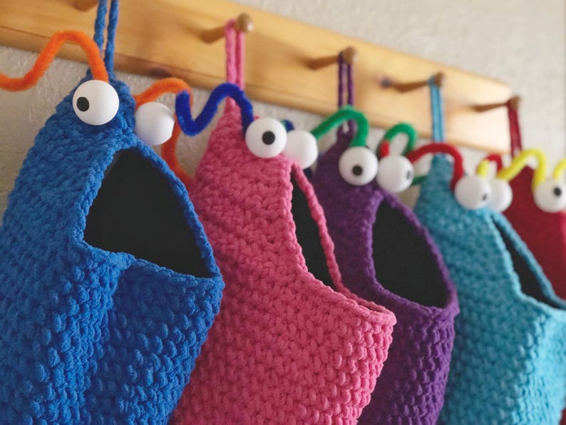 May include: A row of colorful, handmade crochet monster-shaped storage bags hanging on a wooden rack. Each bag is a different color: blue, pink, purple, teal, and red. They have large googly eyes and pipe cleaner antennae.