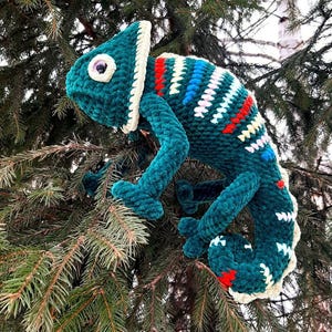 May include: A plush, crocheted chameleon toy featuring a deep teal body with a textured, soft yarn finish. The creature displays a cream-colored crest and underbelly, accented with vibrant stripes of red, white, and blue along its curved back.