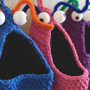 May include: Close-up of several colorful, handmade monster-shaped storage baskets. Each basket is a different color, including blue, pink, purple, and green. They have large googly eyes and pipe cleaner antennae. The baskets have a triangular opening.