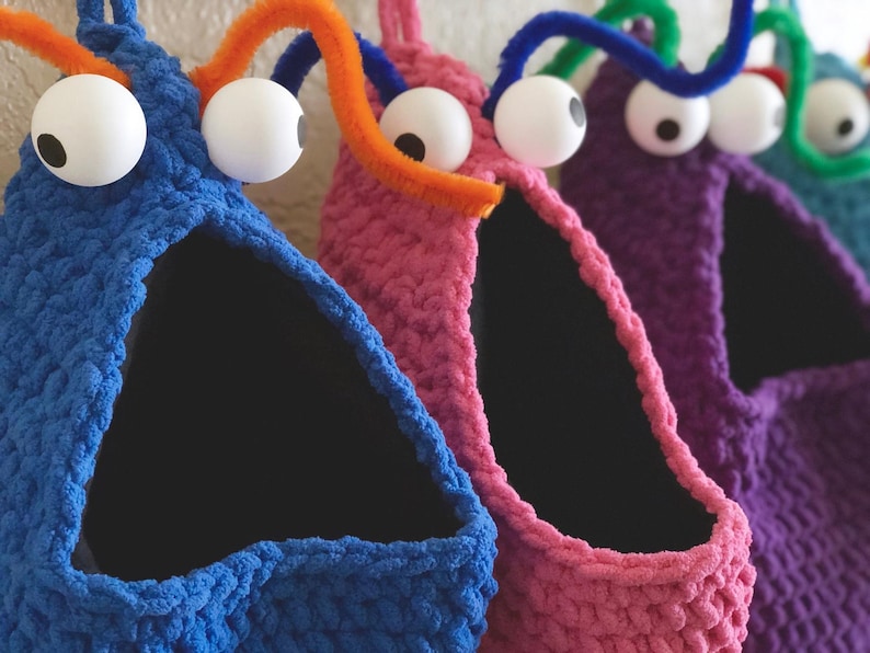 May include: Close-up of several colorful, handmade monster-shaped storage baskets. Each basket is a different color, including blue, pink, purple, and green. They have large googly eyes and pipe cleaner antennae. The baskets have a triangular opening.