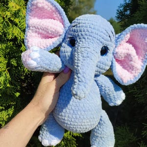 Op de afbeelding: Een handgemaakte blauwe gehaakte olifant knuffel met roze binnenoren en witte accenten op de voeten. De olifant heeft grote, expressieve blauwe ogen en een lange slurf. Het zachte speelgoed wordt omhoog gehouden tegen een achtergrond van groen gebladerte.