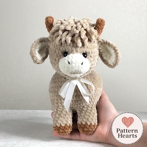 May include: A handmade, plush Highland cow toy in shades of tan, brown, and white. The stuffed animal has a fluffy mane, small horns, and a white bow. The toy is designed with a soft, textured yarn.
