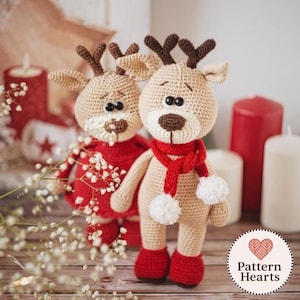 May include: Two handmade crochet reindeer dolls. One reindeer is wearing a red sweater, and the other is wearing a red scarf with white pom-poms. Both have brown antlers, black eyes, and red shoes. The background includes candles and a wooden surface.