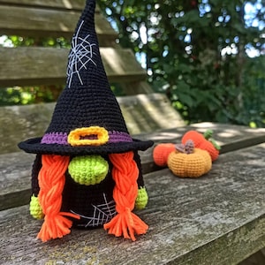 May include: A handmade crochet witch gnome with a black hat, purple band, and orange buckle. The gnome has orange braids, a green nose, and green hands. Small crochet pumpkins are next to the gnome. The gnome is sitting on a wooden bench.