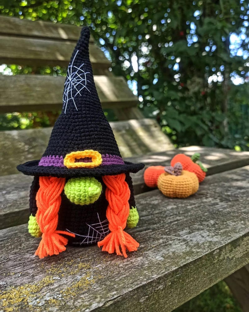 May include: A handmade crochet witch gnome with a black hat, purple band, and orange buckle. The gnome has orange braids, a green nose, and green hands. Small crochet pumpkins are next to the gnome. The gnome is sitting on a wooden bench.
