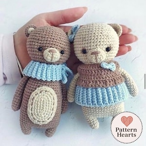 May include: Two handmade crochet teddy bears. One is brown with a light blue scarf, the other cream-coloured with a brown top and blue skirt. Both have black eyes and are holding a heart-shaped logo.