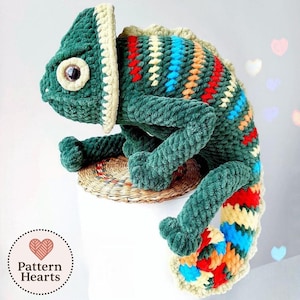 May include: A plush crochet chameleon toy featuring a dark green body with a textured, yarn-based surface. The creature has a cream-colored eye border, a large brown eye, and a tail patterned with stripes of red, yellow, and blue yarn.
