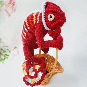 May include: A crocheted chameleon toy in vibrant red yarn with a white trim along its back and head. The creature features a curled tail with multicolored accents and a textured eye. It is perched on a small, light brown woven wicker basket.