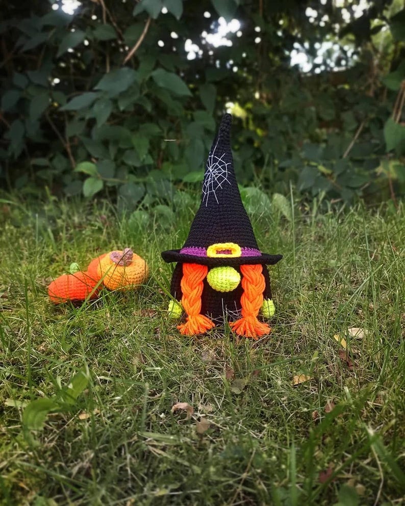 May include: A crocheted Halloween gnome with a black witch hat featuring a spiderweb design, purple brim, and yellow buckle. The gnome has orange braids, green cheeks, and a small orange pumpkin. The gnome is set in a grassy outdoor scene.