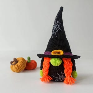 May include: A handmade crochet Halloween gnome with a black witch hat featuring a white spiderweb design. The gnome has orange braids, a green nose, and a purple and gold belt. Two small crochet pumpkins are in the background.