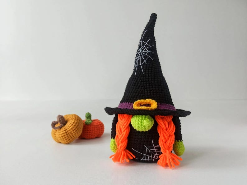 May include: A handmade crochet Halloween gnome with a black witch hat featuring a white spiderweb design. The gnome has orange braids, a green nose, and a purple and gold belt. Two small crochet pumpkins are in the background.