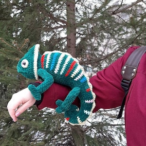 May include: A plush, crocheted chameleon toy featuring a deep green body with a textured surface. The creature is decorated with horizontal stripes in white, red, and blue, and has a large, round eye with a dark pupil and a curled tail.