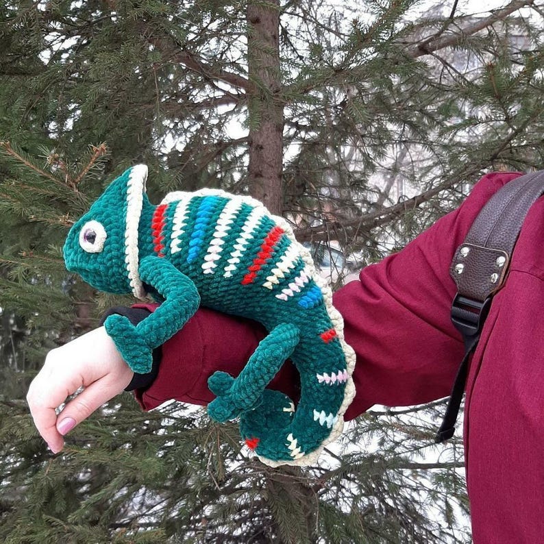 May include: A plush, crocheted chameleon toy featuring a deep green body with a textured surface. The creature is decorated with horizontal stripes in white, red, and blue, and has a large, round eye with a dark pupil and a curled tail.