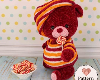 Può includere: Un orsacchiotto all'uncinetto in filato rosso scuro, che indossa un cappello e un maglione a righe arancioni, bianche e rosse. L'orso ha un muso rosa chiaro, occhi neri e tiene in mano un piccolo lecca-lecca a spirale rosso e bianco.