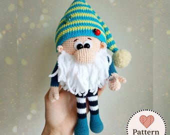 Può includere: Una bambola gnomo all'uncinetto con un cappello alto e appuntito a righe orizzontali verde acqua e gialle, una barba in filato bianco e un naso rotondo beige. La bambola ha gambe a righe bianche e nere, piedi verde acqua e una coccinella rossa.