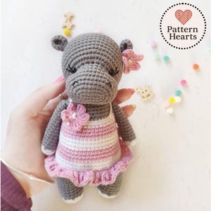 May include: A handmade gray crochet hippo wearing a pink and white striped dress with a pink flower accent. The hippo has small ears and a flower in its hair. The image includes the text "Pattern Hearts".