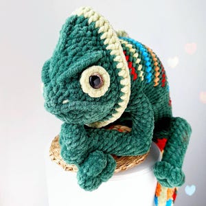 May include: A plush, crocheted chameleon toy crafted from dark green yarn with a cream-colored ridge along its head and back. The creature features a large, dark brown plastic eye and a vibrant, multi-colored striped pattern along its spine.