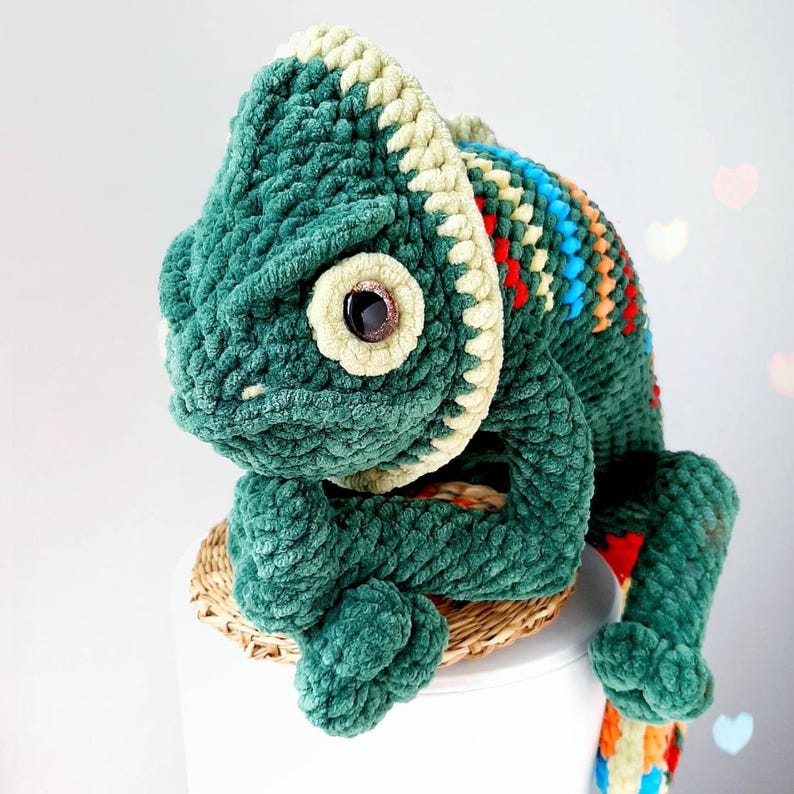 May include: A plush, crocheted chameleon toy crafted from dark green yarn with a cream-colored ridge along its head and back. The creature features a large, dark brown plastic eye and a vibrant, multi-colored striped pattern along its spine.