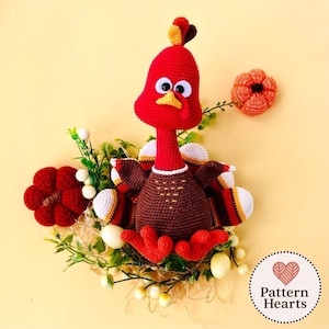 May include: A handmade crochet turkey with a red head, brown body, and white and yellow accents. The turkey is surrounded by greenery, white berries, and two crochet flowers. The image includes a "Pattern Hearts" logo.