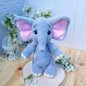 Op de afbeelding: Een handgemaakt, gehaakt olifantenspeelgoed in zacht blauw met roze binnenoren en witte accenten op de voeten. De olifant heeft een slurf, grote oren en blauwe ogen. Het staat op een houten schijf, met witte bloemen op de achtergrond.