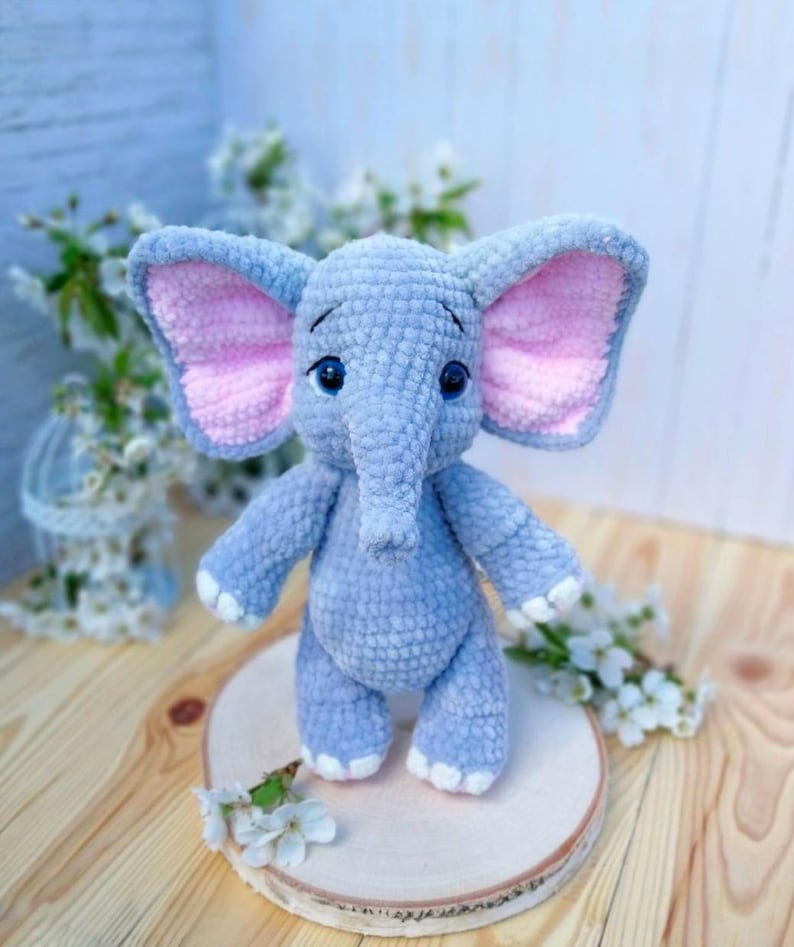 Op de afbeelding: Een handgemaakt, gehaakt olifantenspeelgoed in zacht blauw met roze binnenoren en witte accenten op de voeten. De olifant heeft een slurf, grote oren en blauwe ogen. Het staat op een houten schijf, met witte bloemen op de achtergrond.