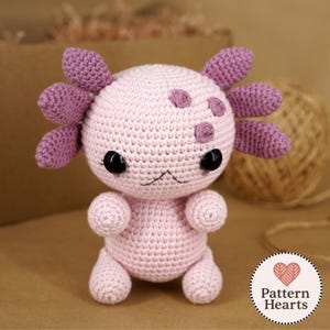 以下が含まれることがあります： 手作りの編みぐるみのウーパールーパーのぬいぐるみ。淡いピンク色で、濃いピンク色のえらと斑点があります。ウーパールーパーは黒い安全な目と縫い付けられた口を持っています。ぬいぐるみは直立しており、小さな腕と脚を見せています。「Pattern Hearts」のロゴが見えます。