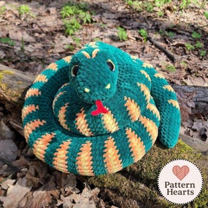 Peut inclure: Un serpent en peluche enroulé en fil vert foncé et orange avec des yeux en plastique noir et une petite langue en feutre rouge. Le serpent présente un aspect texturé et duveteux, disposé en forme circulaire. Un logo indique Pattern Hearts.
