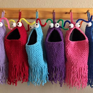 May include: A collection of six handmade, whimsical storage bags in various colors, including purple, red, blue, and pink. Each bag features a large, open mouth design with googly eyes and fringe. They hang from a wooden rack.