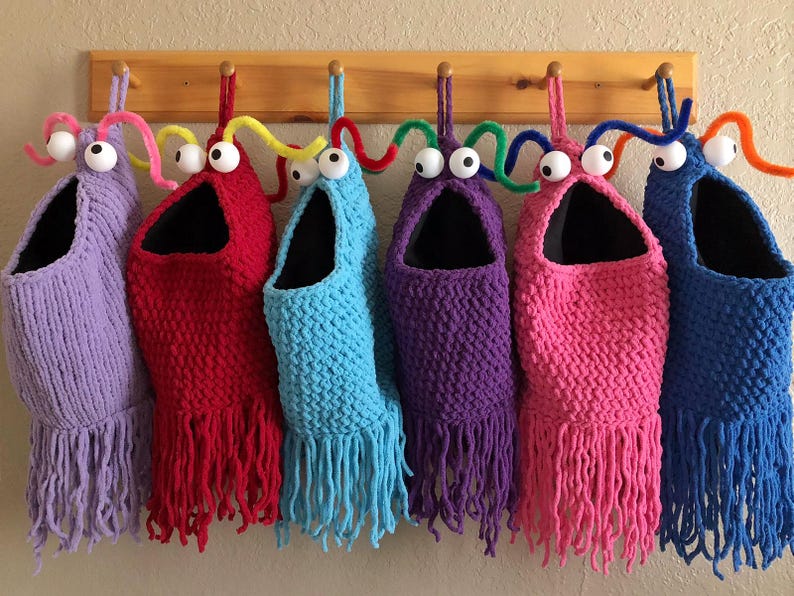 May include: A collection of six handmade, whimsical storage bags in various colors, including purple, red, blue, and pink. Each bag features a large, open mouth design with googly eyes and fringe. They hang from a wooden rack.