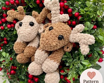 Può includere: Due giocattoli di peluche a forma di renna, nei toni del marrone e beige, con occhi neri e grandi corna, adagiati su un letto di foglie verdi e bacche rosse. Perfetti per le decorazioni natalizie.