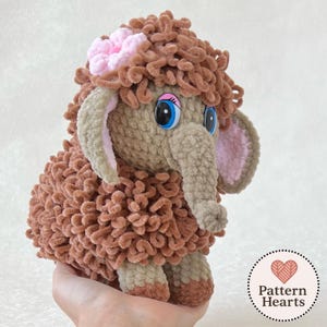 Może przedstawiać: Ręcznie robiona pluszowa zabawka w kształcie mamuta włochatego. Zabawka jest głównie brązowa z puszystą fakturą, dużymi niebieskimi oczami i różowym kwiatkiem na głowie. Uszy i trąba są w jasnobeżowym kolorze.