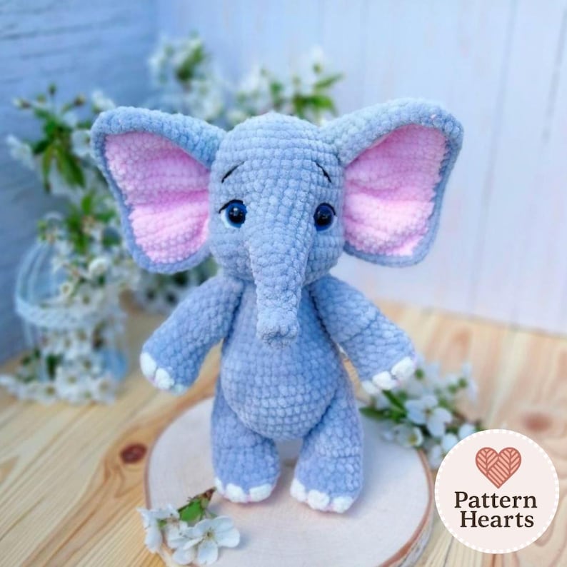 Op de afbeelding: Een handgemaakte, lichtblauwe gehaakte olifant met roze binnenoren en witte accenten op de voeten. De olifant heeft blauwe ogen en een slurf. Het speelgoed wordt gepresenteerd op een houten oppervlak met witte bloemen op de achtergrond.