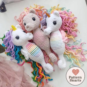 Op de afbeelding: Drie gehaakte zeepaardjes-eenhoorns met regenboogmanen en strepen. De zeepaardjes zijn wit, roze en beige, met gele of oranje hoorns. De manen zijn een mix van regenboogkleuren. De zeepaardjes hebben zwarte knoopogen.