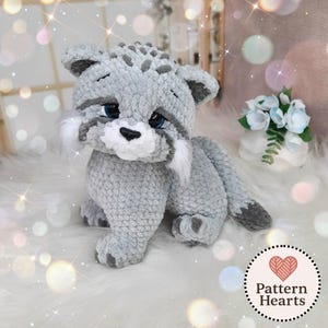 May include: A plush, crocheted stuffed animal resembling a small cat or lynx, crafted from soft, light gray yarn with darker gray spots and white accents on the cheeks. It features large, dark blue eyes, a black nose, and a long, gray tail.