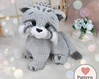 Può includere: Un peluche all'uncinetto che raffigura un piccolo gatto o lince, realizzato in morbido filato grigio chiaro con macchie grigio scuro e accenti bianchi sulle guance. Presenta grandi occhi blu scuro, un naso nero e una lunga coda grigia.